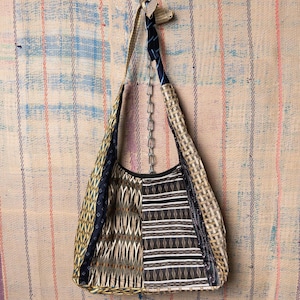 Puede incluir: Bolso de hombro patchwork con una variedad de telas estampadas en tonos beige, negro y azul marino. El bolso tiene una correa larga y está suspendido de una cadena. El fondo es una superficie texturizada y rayada.