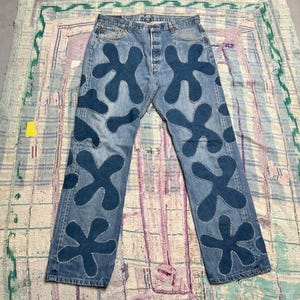 Könnte beinhalten: Hellblaue Jeans aus Denim mit einer Reihe von dunkelblauen, unregelmäßigen, organisch geformten Flicken, die auf die Vorderseite beider Beine genäht sind. Die Hose verfügt über einen Knopfverschluss, Vordertaschen und sichtbare weiße Nähte.