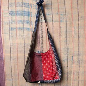 Puede incluir: Un bolso de hombro patchwork hecho de corbatas recicladas. El bolso presenta una variedad de patrones y colores, incluyendo rojo con puntos amarillos, azul marino con puntos blancos y un patrón geométrico gris oscuro. La correa es una corbata azul estampada.