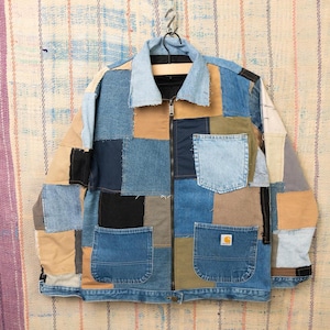 Peut inclure: Veste en jean patchwork composée de carrés de tissu bleus, beiges et noirs. La veste est dotée d'une fermeture éclair, de deux poches avant et d'un petit logo. Elle est suspendue à un cintre.