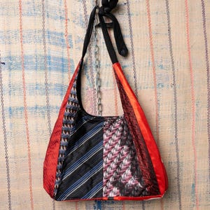 Puede incluir: Un bolso de hombro tipo patchwork hecho con corbatas recicladas. El bolso presenta una variedad de patrones y colores, incluyendo rayas rojas, negras, azules y blancas y diseños geométricos. El bolso tiene una correa negra y una cadena plateada.
