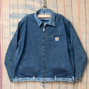 Peut inclure: Veste en jean bleue avec fermeture éclair, col et plusieurs poches. La veste porte le logo Carhartt sur la poitrine et est suspendue à un cintre en bois.