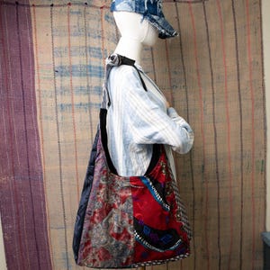 Puede incluir: Un bolso de hombro grande, de retazos, con una correa negra. El bolso presenta paneles de telas rojas, azules y estampadas. El maniquí lleva una camisa a rayas azul claro y una gorra de béisbol vaquera.