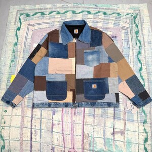 May include: A patchwork jacket constructed from various shades of blue denim and brown canvas fabric. The jacket features a full-length front zipper, a denim collar, long sleeves, and multiple pockets. A small Carhartt brand label is visible.