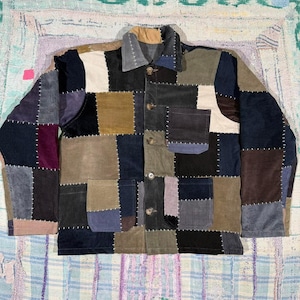 Könnte beinhalten: Eine langärmelige Knopfjacke aus einem Patchwork aus Cordstoffquadraten in verschiedenen Farben wie Marineblau, Beige, Olivgrün, Schwarz und Violett. Das Kleidungsstück verfügt über einen klassischen Kragen, eine Knopfleiste vorne und zwei Taschen.