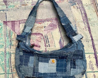 May include: A hobo-style shoulder bag constructed from a patchwork of various blue denim fabric squares. The bag features a single integrated shoulder strap and a small rectangular Carhartt brand patch centered on the front exterior panel.