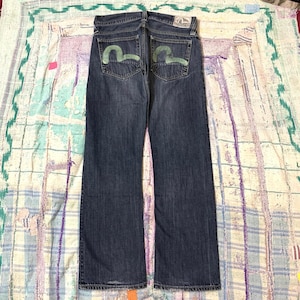 Puede incluir: Vaqueros de mezclilla azul oscuro con un lavado desgastado y corte recto. Los bolsillos traseros están adornados con un logotipo de gaviota pintado a mano en verde claro. Un parche blanco tipo cuero con texto negro está cosido en la cintura.