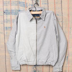 Peut inclure: Veste zippée gris clair avec col boutonné et manches longues. La veste présente un petit logo brodé sur la poitrine et est suspendue à un cintre noir. La veste est en tissu léger.