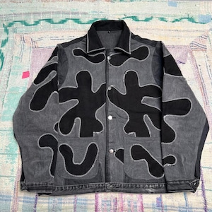 Peut inclure: Une veste en jean gris anthracite ornée de pièces de tissu noir aux formes organiques, cousues sur le devant et les manches avec une broderie en zigzag blanche. La veste présente un col classique, une fermeture boutonnée et deux poches.