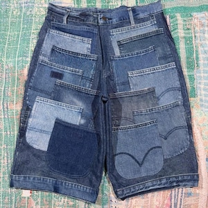 May include: Denim shorts featuring a patchwork design composed of multiple rectangular and square fabric panels in varying shades of blue. The garment includes a waistband with belt loops, a button closure, and several visible pocket shapes.