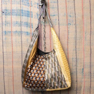 Puede incluir: Un bolso tote hecho a mano con un diseño de patchwork. El bolso presenta una variedad de telas en tonos dorados, marrones, grises y azules. El bolso tiene una correa larga y un detalle de cadena. El bolso está colgado contra un fondo a rayas.