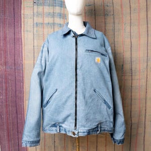 Peut inclure: Veste en jean bleu clair avec une fermeture éclair noire, deux poches avant et une petite poche poitrine. La veste a un col et un petit écusson jaune et blanc sur la poitrine. La veste est présentée sur un mannequin.