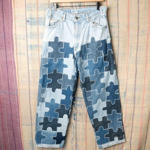 Peut inclure: Jean clair avec un design patchwork unique. Le jean présente des pièces de puzzle de différentes nuances de bleu, créant un effet visuel saisissant. Le jean est suspendu à un cintre en bois.