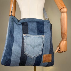 May include: A shoulder bag constructed from vertical strips of blue denim in varying shades. The bag features a single external patch pocket made from light blue denim and a rectangular brown leather-like brand patch in the bottom right corner.