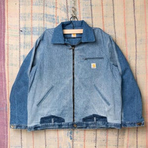 Peut inclure: Veste en jean avec fermeture éclair, corps bleu clair et manches et col bleu foncé. La veste a deux poches avant, une petite poche poitrine et le logo Carhartt.