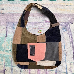 May include: A hobo-style shoulder bag constructed from patchwork corduroy fabric in various colors including navy, olive, tan, grey, and cream. The bag features a single wide strap and a prominent, rectangular pink corduroy pocket on the front.