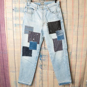 May include: Light wash denim jeans with various fabric patches in shades of blue, black, and striped patterns. The jeans have a straight leg fit and are lightly distressed with white paint splatters.