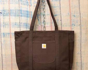 May include: A brown tote bag with multiple pockets and long handles, suspended from a metal chain. The bag features a small, square logo patch on the front. The bag is made of a durable material, suitable for carrying various items.