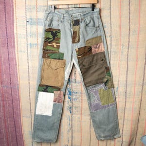 May include: Light blue denim jeans with multiple patches in various colors and patterns, including camouflage and solid fabrics. The patches are sewn onto the jeans, creating a unique, repurposed design. The jeans are hanging on a wooden hanger.
