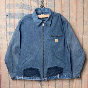 Peut inclure: Veste en jean bleue avec fermeture éclair, col chemise et plusieurs poches. La veste présente un petit écusson carré sur la poitrine et deux empiècements en jean plus foncés en bas. La veste est suspendue à un crochet en métal.