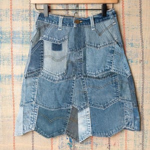 May include: A patchwork denim skirt constructed from various shades of blue jean fabric. The skirt features multiple pockets, visible seams, and a high-waisted design with a metal button closure. The hemline is uneven, showcasing different denim textures and washes.