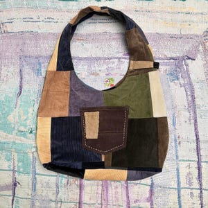 May include: A hobo-style shoulder bag constructed from patchwork corduroy fabric in various colors including olive green, navy blue, brown, tan, and cream. The bag features a central dark brown exterior pocket with visible light-colored stitching.