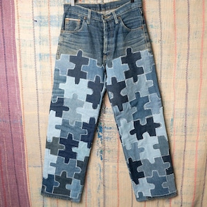 May include: Blue denim jeans with puzzle piece patchwork in various shades of blue. The patchwork covers the lower portion of the jeans, creating a unique and artistic design. The jeans are hanging on a hanger.