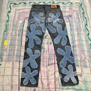 Può includere: Jeans in denim scuro con grandi toppe in tessuto azzurro a forma di fiore cucite sul retro e sulle gambe. I jeans includono un'etichetta del marchio in pelle marrone chiaro sulla cintura posteriore, due tasche posteriori e cuciture bianche.
