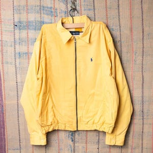 Peut inclure: Veste jaune à fermeture éclair avec col et petit logo bleu marine sur la poitrine. La veste a des manches longues et des poignets et un ourlet côtelés. La veste est suspendue à un cintre en bois.