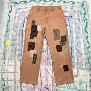 May include: Tan canvas work pants featuring multiple rectangular patches in various shades of brown, olive, and cream. The patches are secured with visible white sashiko-style stitching, creating a textured, reinforced appearance on the legs.