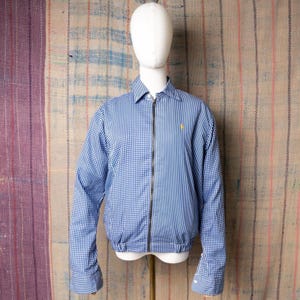 Peut inclure: Veste bleue et blanche au design unique. Le côté gauche présente un motif vichy, tandis que le côté droit arbore des rayures verticales. Un petit logo jaune est visible sur la poitrine droite. La veste est zippée et a un col.