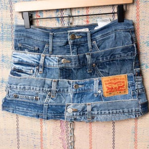 May include: A patchwork denim skirt made from repurposed jeans. The skirt features various shades of blue denim, with visible pockets, belt loops, and metal button closures. A red Levi Strauss & Co. label is sewn on the front. The skirt is hanging on a wooden hanger.