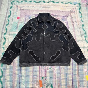 May include: A black denim jacket featuring a fold-down collar, long sleeves, and a front button closure. The jacket is decorated with large, abstract, amoeba-like shapes outlined in thick, white, decorative zigzag stitching.