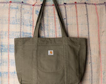 May include: Olive green canvas tote bag with a rectangular front pocket and two side pockets. The bag has long shoulder straps and a small, square logo patch on the front pocket. The bag is hanging against a patterned fabric background.