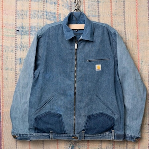 Peut inclure: Veste en jean bleue avec une fermeture éclair intégrale, une petite poche poitrine et deux poches avant. La veste présente un col et l'écusson du logo Carhartt. La veste est suspendue à un crochet en métal.