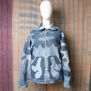 Peut inclure: Veste en jean bleu clair avec un design unique. La veste présente un motif patchwork de formes bleu clair, créant un look distinctif. Elle a un col classique, une fermeture boutonnée et deux poches avant. La veste est présentée sur un mannequin.