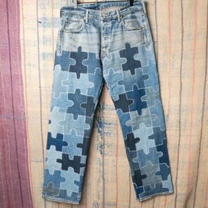 May include: Light blue denim jeans with a unique patchwork design. The jeans feature puzzle piece-shaped patches in various shades of blue, creating a visually striking pattern. The jeans have a classic five-pocket design and a button fly.