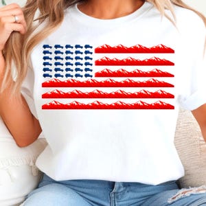 Könnte beinhalten: Ein weißes T-Shirt mit einem Grafikdesign, das an die amerikanische Flagge erinnert. Das obere linke Feld besteht aus kleinen blauen Geländewagensilhouetten, während die Streifen aus sich wiederholenden roten Bergketten mit weißen Gipfeln bestehen.