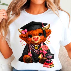 May include: White t-shirt featuring a cartoon Highland cow wearing a graduation cap and gown. The cow holds a bouquet and a sign that says "Congrats Grad." The design includes the text "Class of 2026."