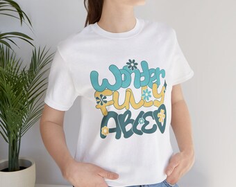 May include: A white short-sleeve t-shirt featuring the text "WONDERFULLY ABLED" in a layered, multi-colored graphic design. The lettering is in shades of teal, yellow, and dark green, accented with small flower icons and a heart shape.