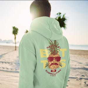 Puede incluir: Una sudadera con capucha verde claro con un gráfico de piña en la espalda. La piña lleva gafas de sol y un bigote. El texto "VIBES" está debajo de la piña. El texto "SUMMER TIME" está encima de la piña. El texto "BUT" está a ambos lados de la piña.