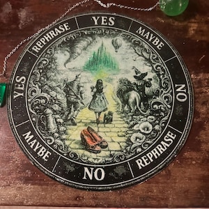 May include: Round black and white decorative planchette with a chain and a green crystal pendulum. The planchette features an illustration of the Wizard of Oz characters and the words "Yes", "Maybe", "No", and "Rephrase".