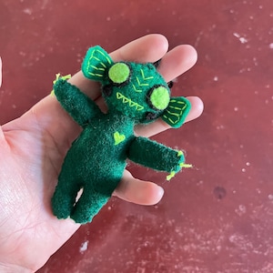 Può includere: Una creatura fatta a mano in feltro verde con grandi occhi rotondi e piccole corna. La creatura ha un disegno cucito sulla testa e sul corpo, con accenti di filo giallo sugli arti. Il giocattolo è tenuto in una mano.