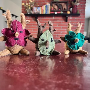 Könnte beinhalten: Drei handgefertigte Filzfiguren mit blüten- und blattförmigen Gesichtern und geweihartigen Geweihen. Die Figuren haben braune Körper und sind auf einer roten Oberfläche angeordnet. Eine hat ein magentafarbenes Blumengesicht, eine ein grünes Blattgesicht und eine ein türkisfarbenes Blumengesicht.