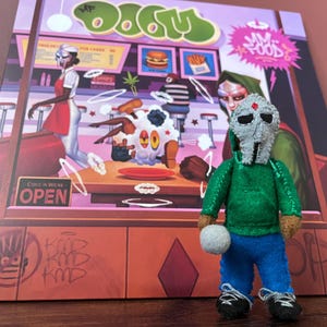 May include: A handmade felt doll of a character wearing a silver mask, green hoodie, blue trousers, and black trainers. The doll is in front of a colourful album cover with the word "DOOM" and a diner scene.