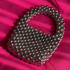 Peut inclure: Un sac à main en perles argentées de forme ronde. Le sac est composé de nombreuses petites perles rondes tissées ensemble.