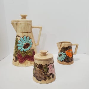 Puede incluir: Un juego de café de cerámica beige con diseños florales y frutales. El juego incluye una cafetera alta, una pequeña crema y un azucarero. Las decoraciones presentan flores azules, fresas y una calabaza naranja.