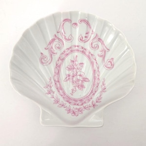 May include: A white ceramic dish shaped like a scallop shell, featuring a pink transferware-style print. The design includes a central oval frame containing a floral motif, surrounded by decorative scrollwork and a leafy border at the base.