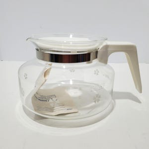 May include: Clear glass coffee carafe with a white lid and handle. The carafe has a silver band around the middle and a floral pattern. A paper tag is inside the carafe.