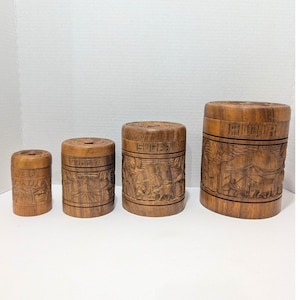 Puede incluir: Un juego de cuatro recipientes cilíndricos de madera de distintos tamaños, cada uno con escenas de paisajes talladas y etiquetas que dicen TEA, COFFEE, SUGAR y FLOUR. Los recipientes tienen tapas a juego y un acabado de veta de madera natural.